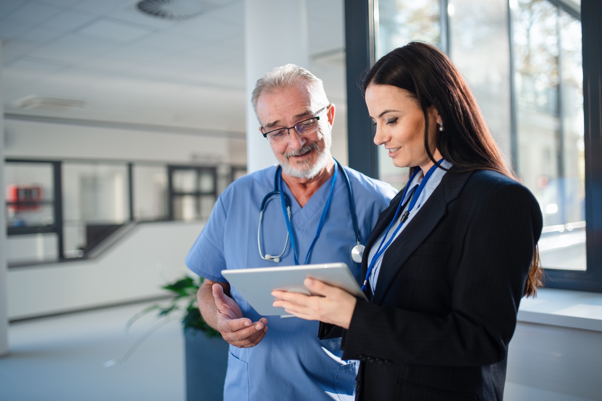 A senior physician in scrubs and a healthcare administrator review information together on a tablet in a bright hospital hallway, representing the credentialing and provider management professionals MD-Staff serves.