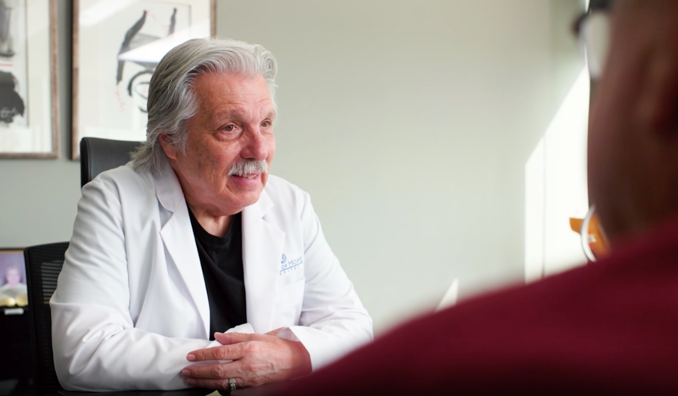 Dr. Contreras in a white Oasis of Hope lab coat seated at a desk in conversation with a patient across from him, representing the doctor-patient consultation experience that the rebuilt website needed to communicate clearly to prospective patients and families.