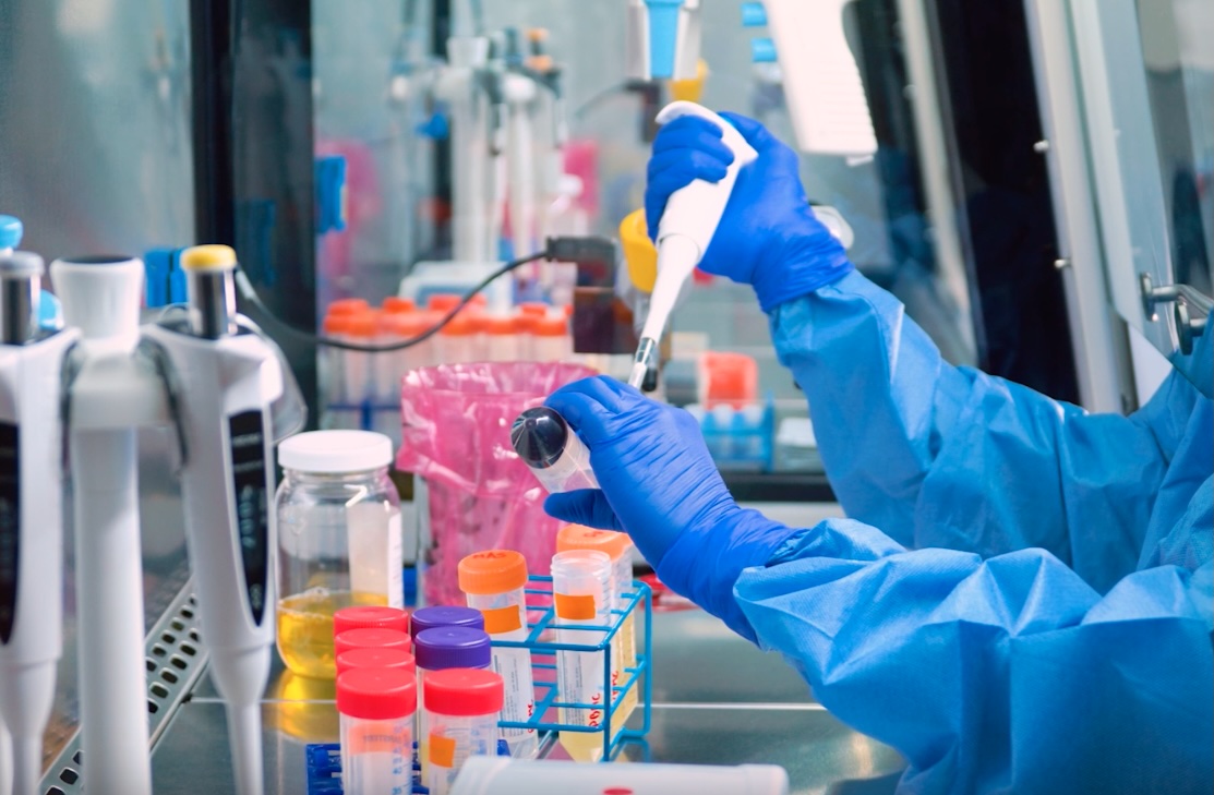 A researcher in full blue PPE and gloves uses a precision pipette in a clinical laboratory surrounded by sample vials and reagents, representing the IMPACT immunotherapy program that received its own dedicated landing page in the Oasis of Hope platform rebuild.