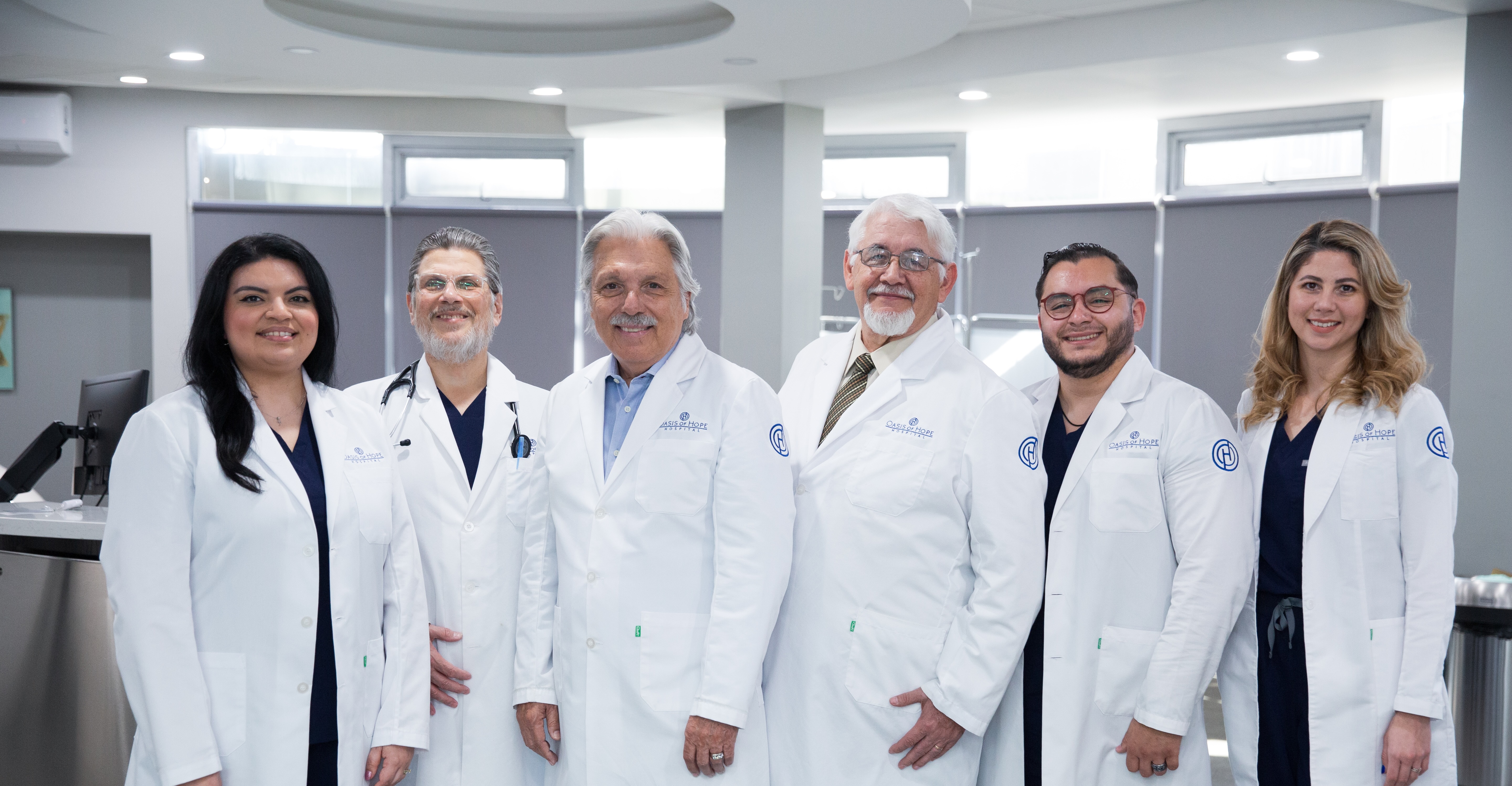 Six Oasis of Hope Hospital physicians and medical specialists in white lab coats with the Oasis of Hope logo standing together in a modern clinical setting, representing the 30-doctor roster given dedicated profile pages on the rebuilt WordPress platform.