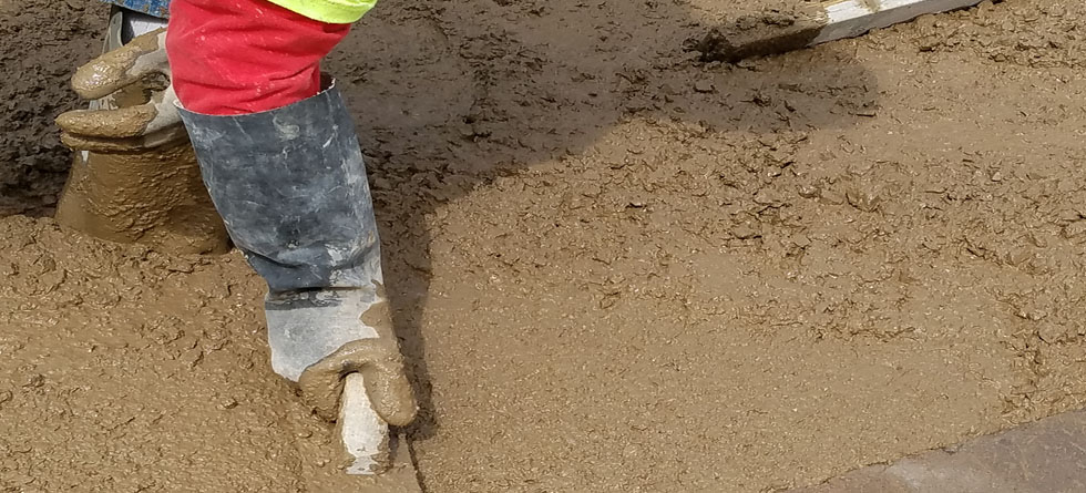 A construction worker in safety gear kneels to hand-smooth freshly poured wet concrete with a trowel on a jobsite, showing the hands-on quality of Superior Ready Mix's ready mix concrete delivery and finishing work.