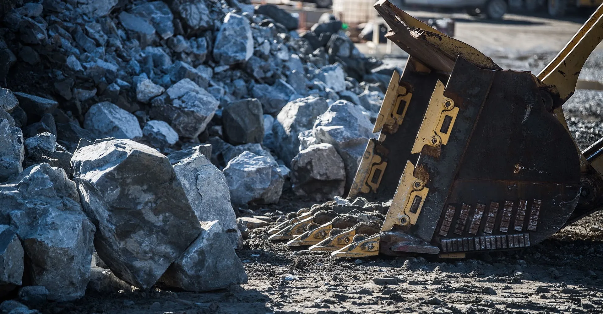An excavator bucket lifts aggregate rock from a large pile at a construction materials quarry site, representing the aggregates and materials supply side of Superior Ready Mix's business.