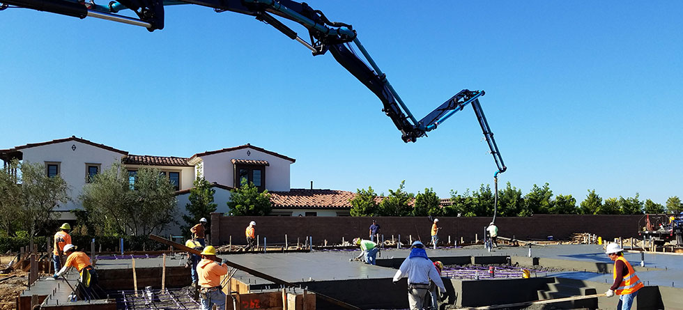 A concrete pump boom extends over a residential construction site in Southern California where a crew of workers in hard hats and safety gear spreads freshly poured ready mix concrete across a reinforced slab foundation.