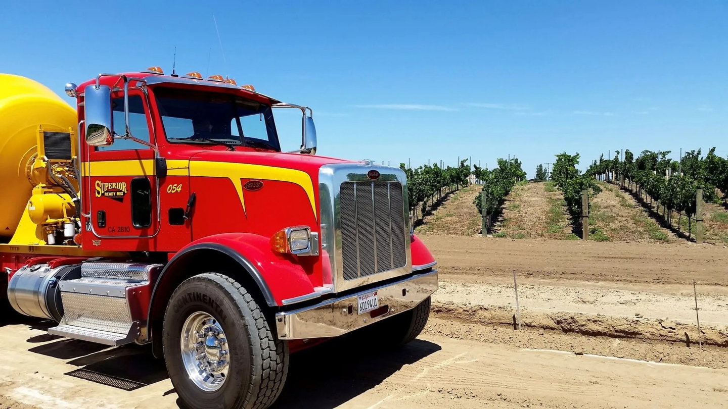 A red and yellow Superior Ready Mix concrete truck parked in front of rows of vines in a California agricultural field under a clear blue sky, showing the fleet's reach across Southern California's diverse project landscape.