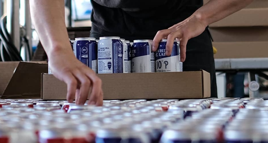 Hands packing White Labs branded beer cans into a cardboard box on a production line, showing the From Cell to Sip brewing program that generates over 300 documented batches of on-site content for the platform.