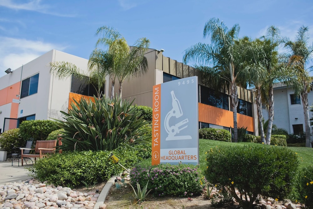 Exterior of the White Labs Global Headquarters building in San Diego, featuring the Tasting Room signage with a microscope icon and the address 9495, set against a clear blue sky with palm trees — representing the brand's California home and physical presence.