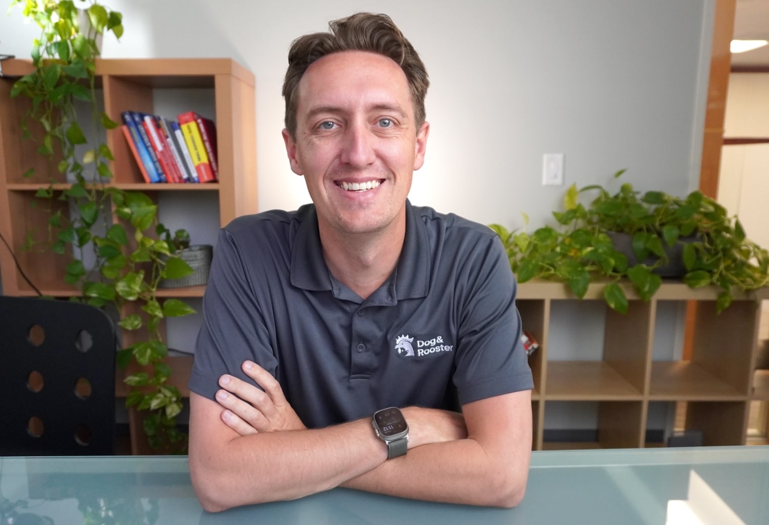 Professional portrait of Kyle Hennessy, Founder and Principal of Dog and Rooster, wearing a grey polo with the company logo, seated at a desk in a bright office.